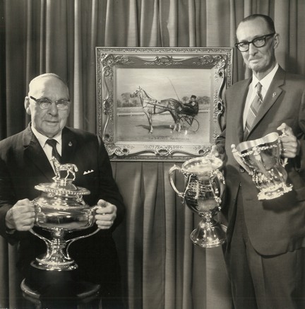 Black and White Photo showing two older men holding trophies standing in front of a painting