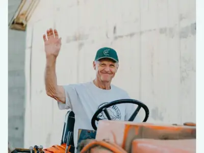 Persson sitting behind the wheel of a tractor waving at the camera.